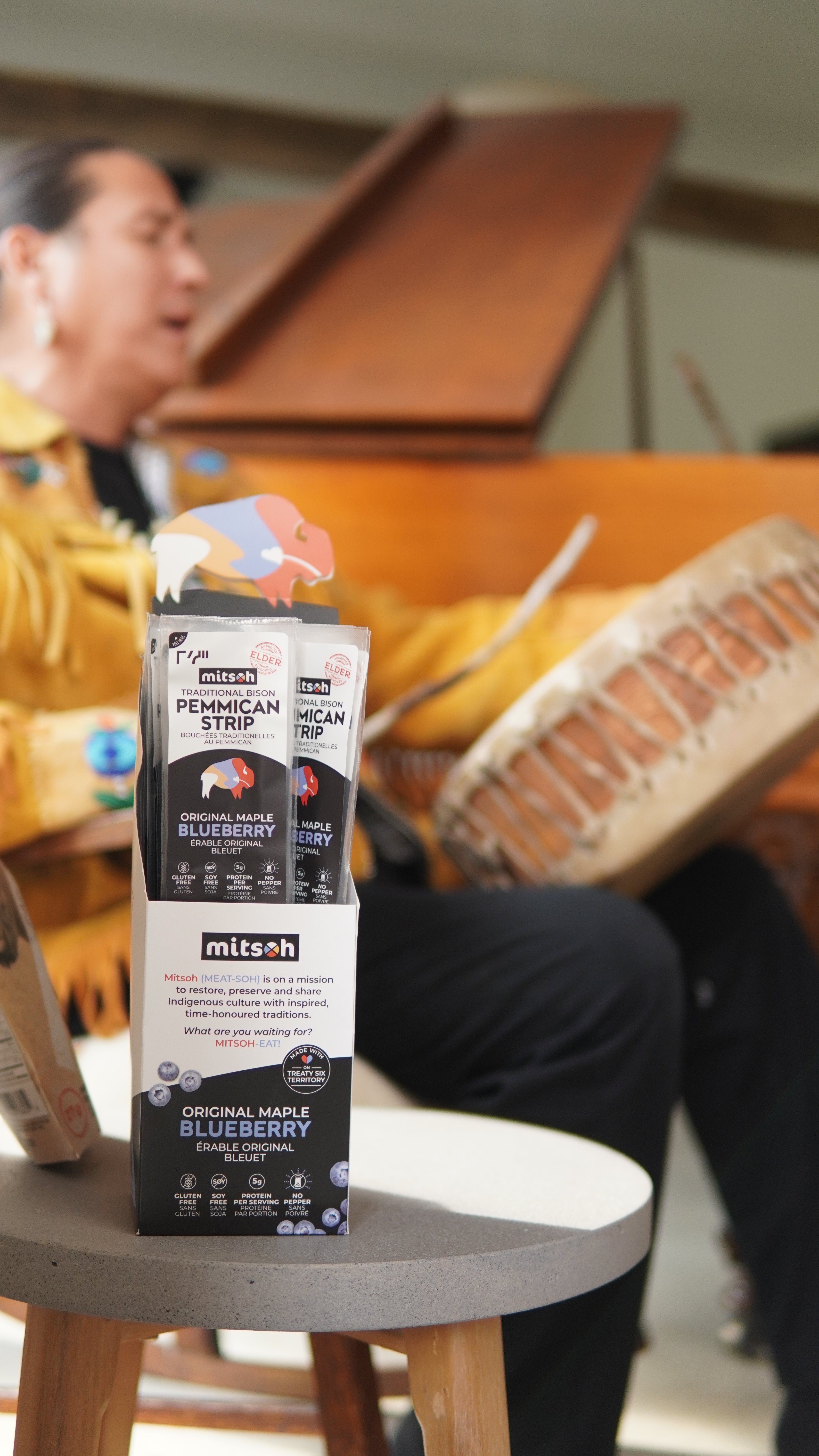 Mitsoh pemmican strips displayed in a retail carton, with founder Ian Gladue seated in the background holding a drum