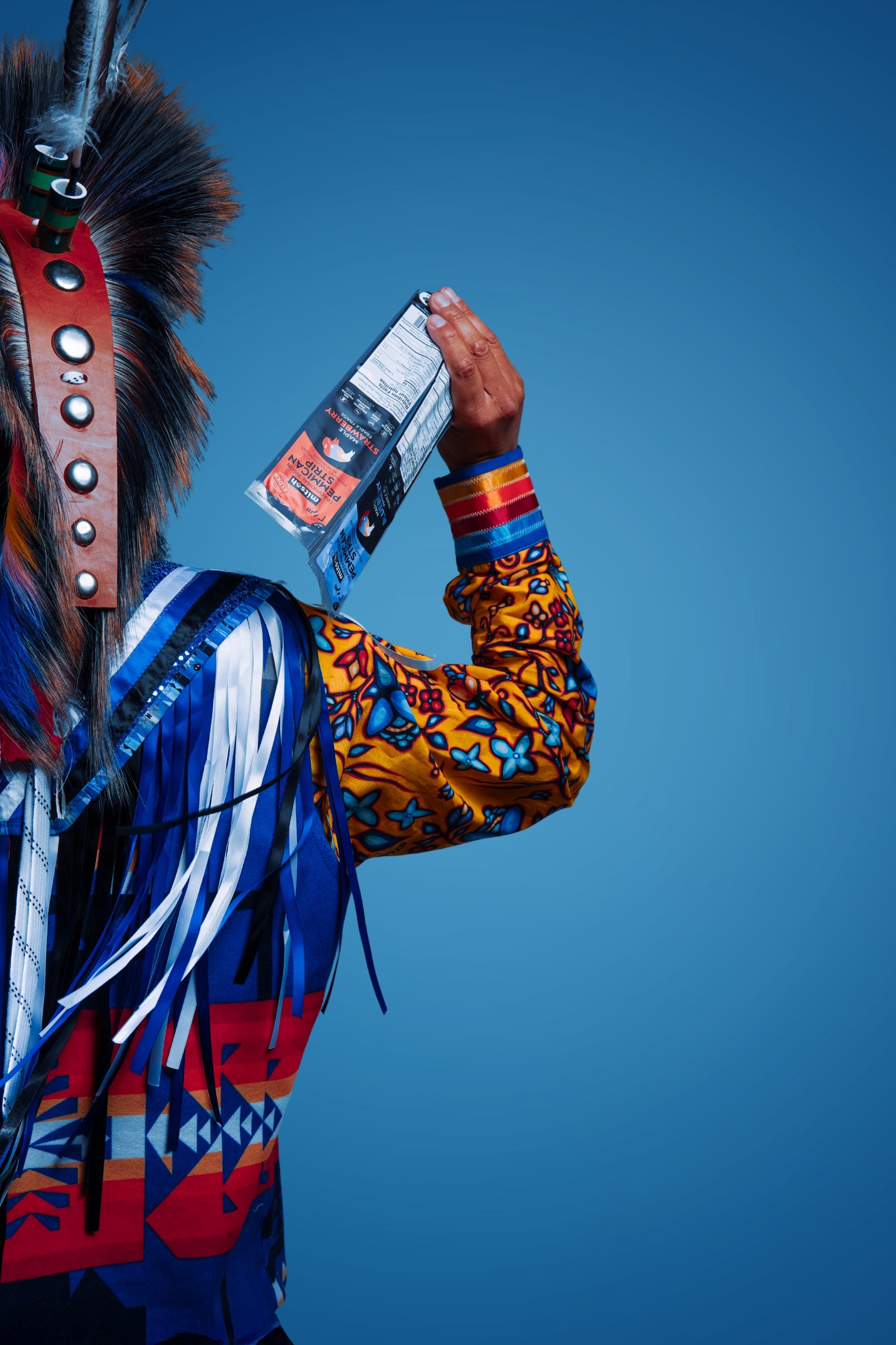 Mitsoh founder Ian Gladue in traditional regalia holding a Mitsoh bison snack against a blue background