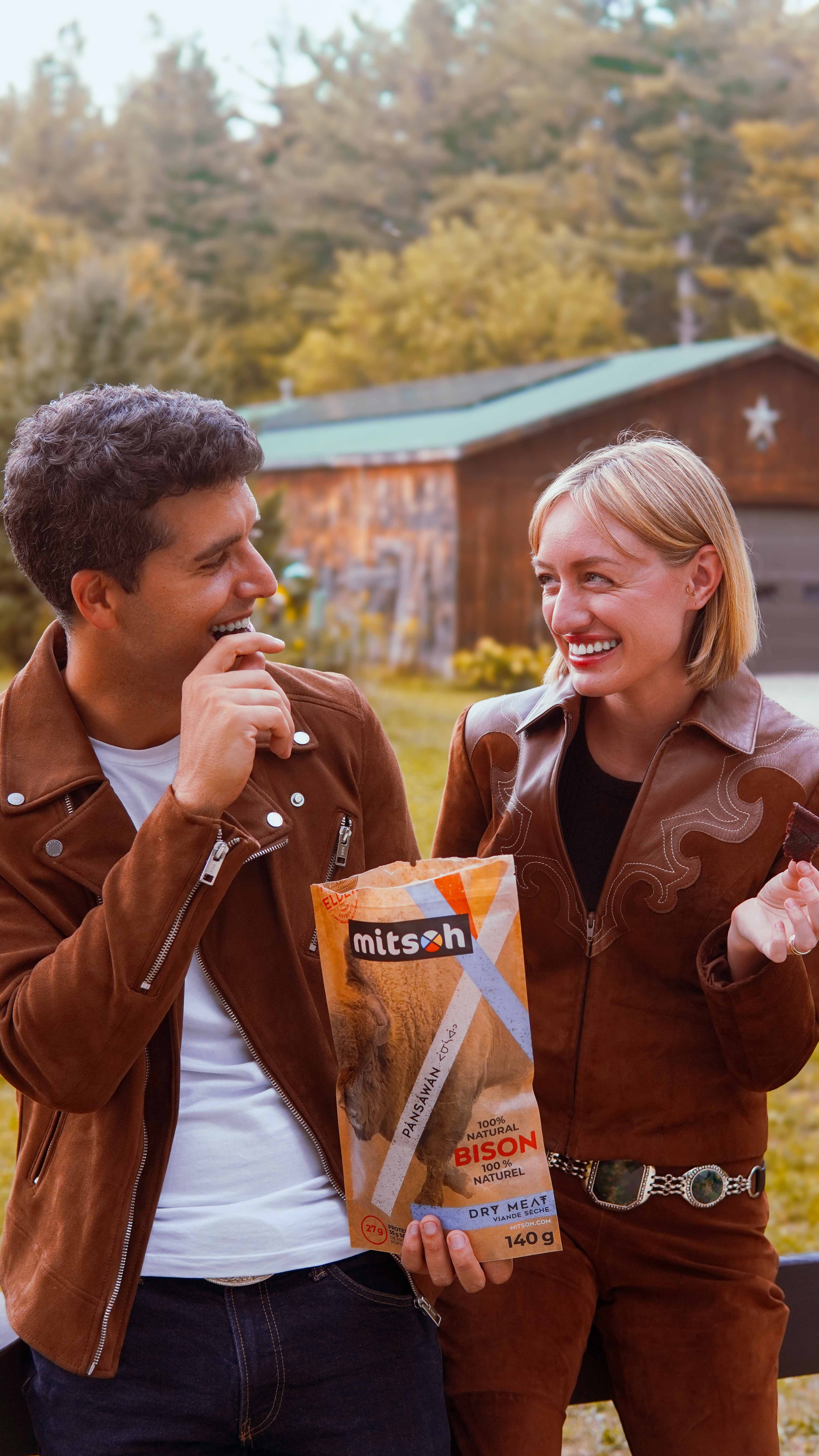 Two people smiling outdoors while sharing a package of Mitsoh dry bison meat