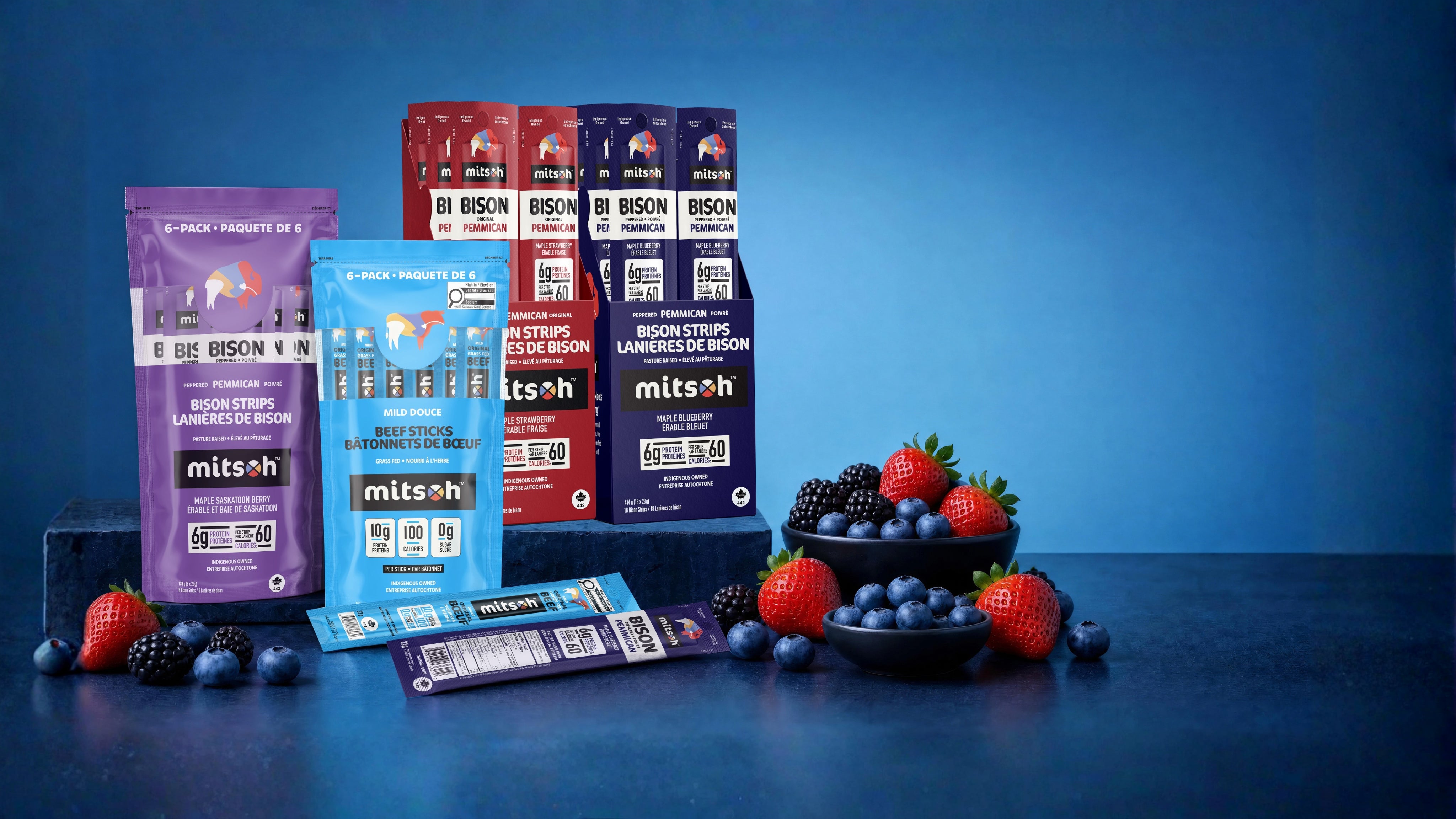 Mitsoh bison pemmican strips and grass-fed beef stick products displayed with berries against a blue studio background
