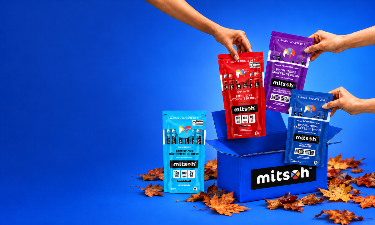 Mitsoh beef stick and bison pemmican packs held above a blue gift box, with autumn leaves and a deep blue background.