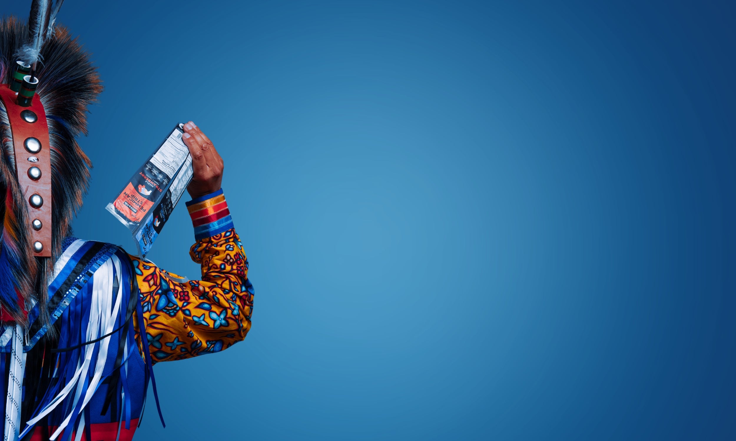 Mitsoh founder Ian Gladue in traditional regalia holding a Mitsoh bison snack against a blue background