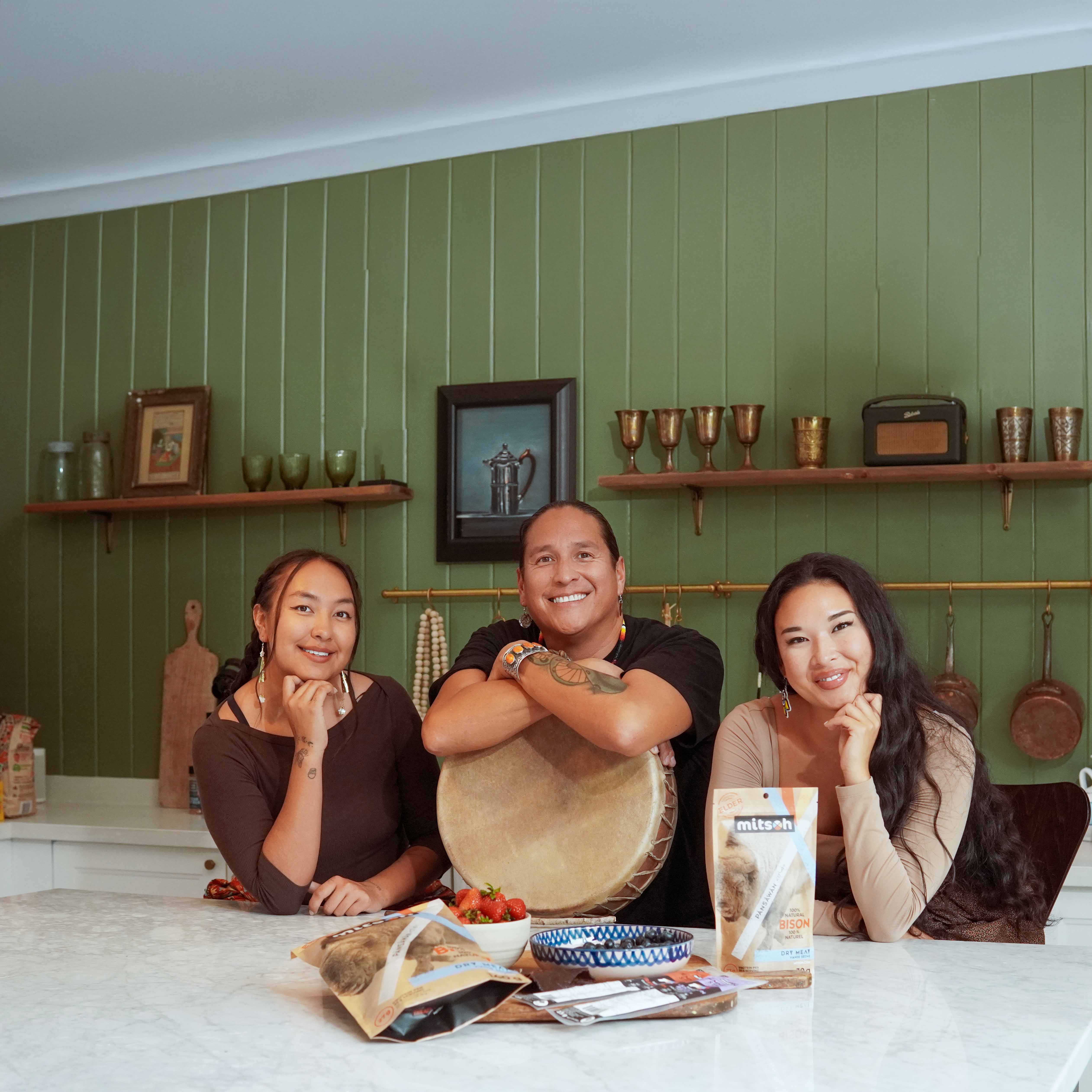 Mitsoh founder Ian Gladue holding a traditional hand drum with two family members gathered around Mitsoh bison snacks