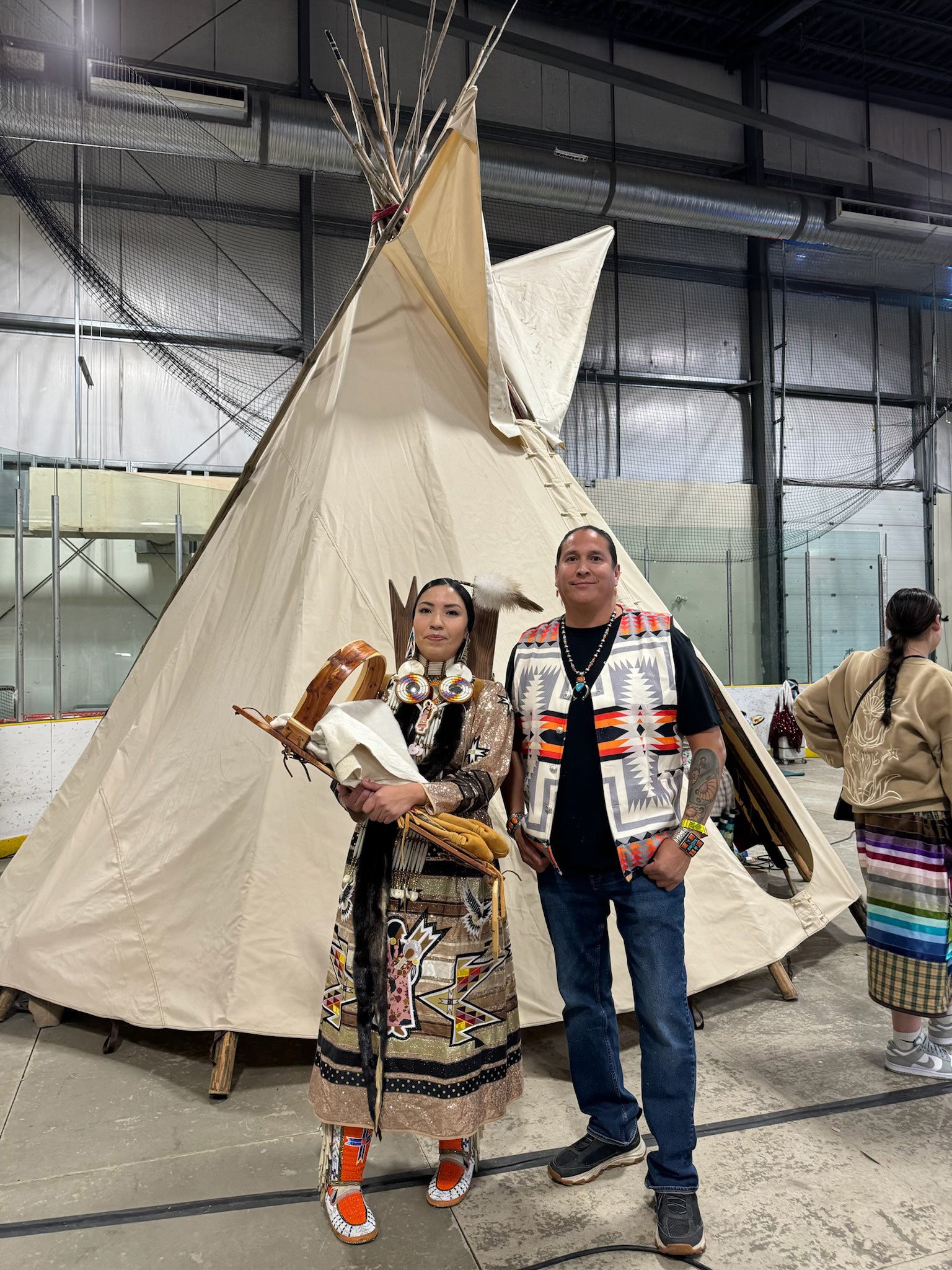 Mitsoh founder Ian Gladue standing with a woman in traditional regalia beside a tipi